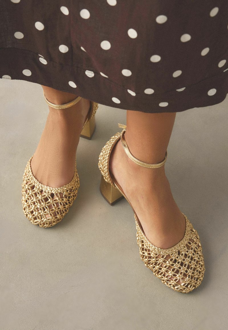 Pieds portant des sandales dorées tissées à talons blocs avec des brides à la cheville, sous une jupe marron à gros pois blancs.
