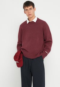Suéter burdeos con cuello acanalado y puños, llevado sobre una camisa blanca con cuello. Se sostiene una gorra roja con la mano izquierda, combinada con pantalones a rayas.
