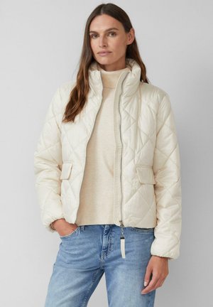 Giacca imbottita crema con colletto alto, zip frontale e due tasche laterali. Indossata sopra un maglione beige e abbinata a jeans blu.