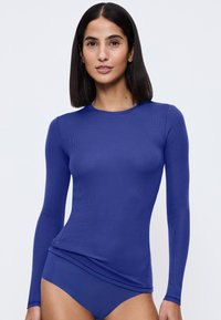 Vrouw met stijlvol zwart haar in een aangesloten lange mouw blauwe top en bijpassend blauwe ondergoed tegen een lichtgrijze achtergrond.