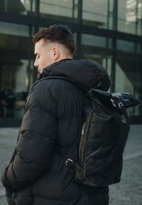 Black leather backpack with a roll-top design, featuring a zippered pocket and adjustable strap, worn over a black puffer jacket.