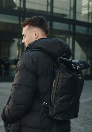 Black leather backpack with a roll-top design, featuring a zippered pocket and adjustable strap, worn over a black puffer jacket.