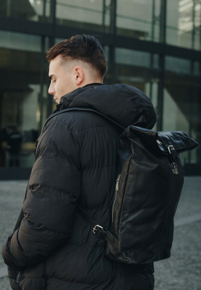 Black leather backpack with a roll-top design, featuring a zippered pocket and adjustable strap, worn over a black puffer jacket.