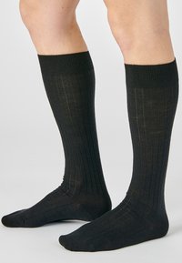 Gambe che indossano calze nere a coste fin sotto al ginocchio, poggianti su una superficie grigia chiara con uno sfondo semplice e chiaro.