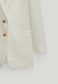 Klasyczna lniana marynarka w kolorze ecru, z dwiema brązowymi guzikami, pojedynczą kieszenią na piersi oraz wyraźną sylwetką z prostymi liniami.