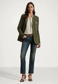 Γυναίκα φορεί φανελένιο πράσινο σακάκι, λευκό πουκάμισο, μπλε τζιν, καστανή ζώνη και καφετί ψηλοτάκουνες σανδάλια, ενώ στέκεται με φόντο απλό.