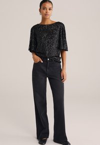 Top nero con paillettes e maniche corte abbinato a jeans neri a vita alta. L'outfit presenta una vestibilità rilassata e dettagli delicati.