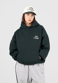 Donna che indossa un ampio felpa verde scuro e un berretto beige, entrambi con il logo "Prohibited 10119 Berlin", in piedi con le mani nella tasca del felpa.