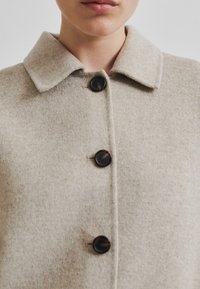 Béžový vlněný kabát s límcem, pěti tmavými knoflíky a texturovanou tkaninou s jemnými skvrnami. Detailní pohled na přední část.