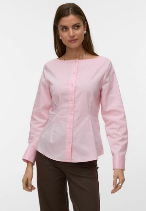 Femme portant un chemisier rose clair boutonné à manches longues et un pantalon marron foncé, posant avec un léger sourire.