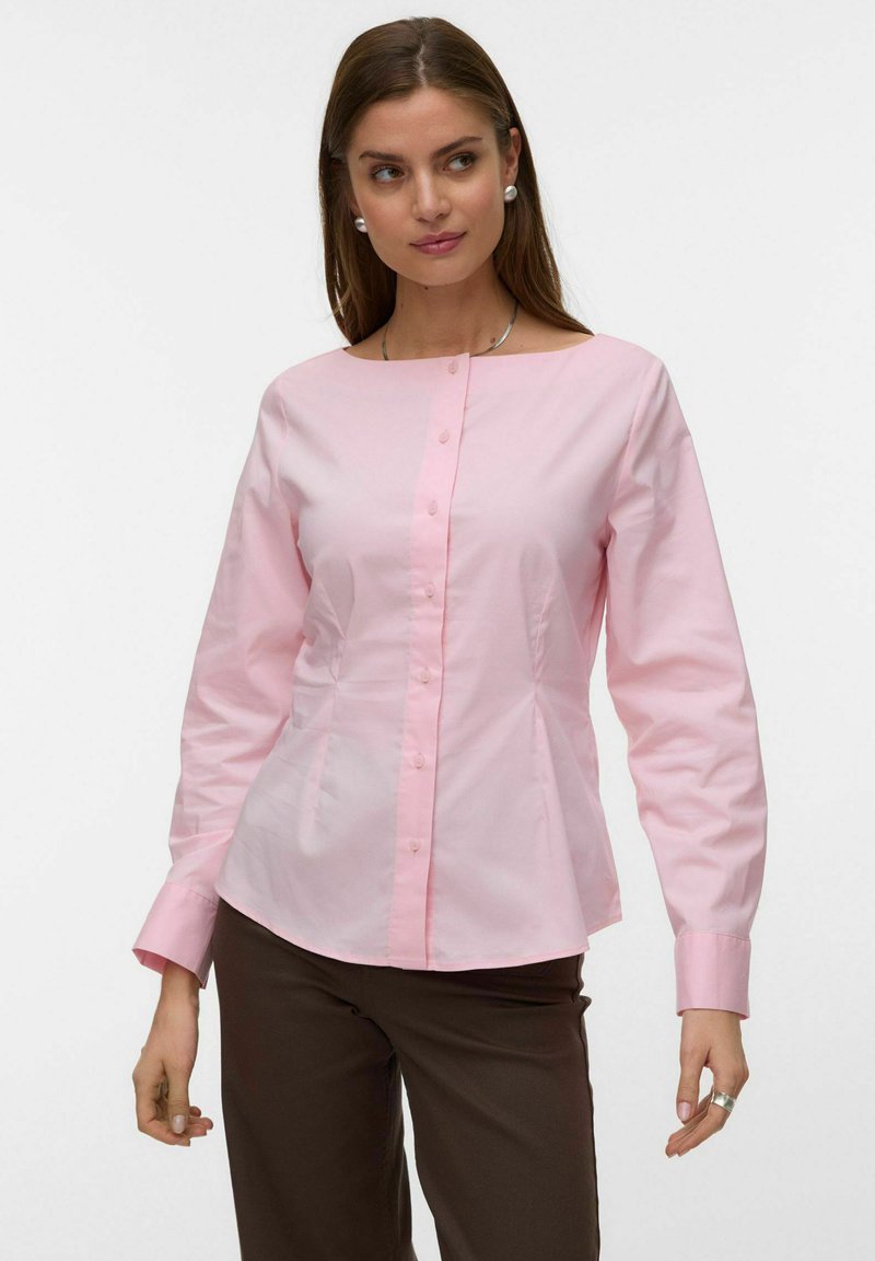 Femme portant un chemisier rose clair boutonné à manches longues et un pantalon marron foncé, posant avec un léger sourire.