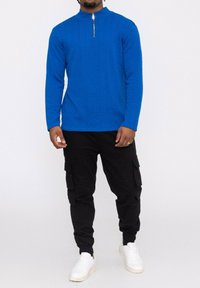 T-shirt bleu en tricot à manches longues avec une demi-fermeture éclair, associé à un pantalon cargo noir et des baskets blanches. Texture lisse et style ajusté.
