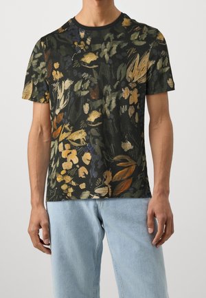 Mann trägt schwarzes T-Shirt mit kurzen Ärmeln und mehrfarbigem, abstraktem Blumenmuster sowie hellblaue Jeans, steht vor einfarbigem Hintergrund.