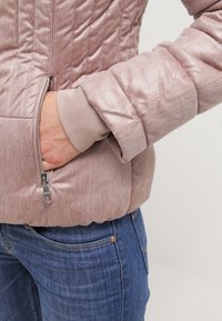 Main dans la poche d'une veste rose texturée avec une fermeture éclair argentée, portant un jean en denim bleu, sur un fond clair uni.