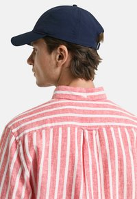 Gestreiftes Hemd in Rot und Weiß mit einem button-down Kragen, das auf der Rückseite eine Falte hat. Eine marineblaue Mütze rundet das Outfit ab.