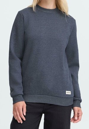 Femme portant un sweat-shirt uni gris foncé à manches longues avec une petite étiquette blanche "oxmo" près de l'ourlet, associé à un pantalon noir.