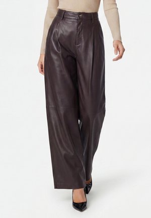 Donna con pantaloni di pelle marrone scuro a gamba larga, top beige a coste a maniche lunghe e tacchi neri a punta, in piedi su sfondo bianco.