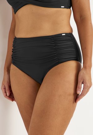 Kvinde iført ruched sorte bikinitrusser med høj talje og matchende badetops, stående med den ene hånd afslappet ved siden.