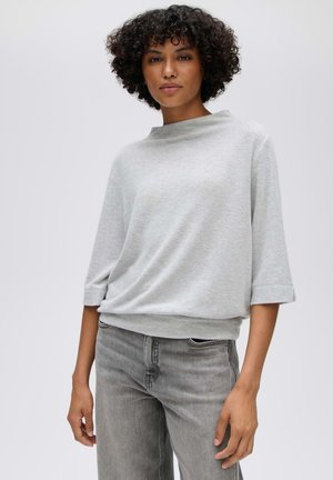 Vrouw met kort krullend haar die een lichtgrijze losse top met driekwartmouwen en vervaagde grijze spijkerbroek draagt tegen een effen achtergrond.