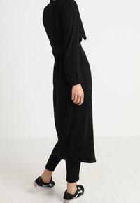 Osoba ubrana w długi czarny płaszcz, na czarne spodnie i czarne sneakersy z białymi podeszwami, stojąca na tle jednolitego białego tła.