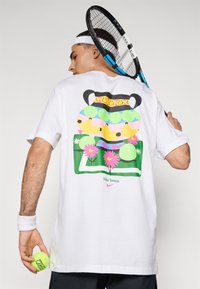 Camiseta blanca de algodón con un diseño gráfico colorido que presenta motivos de tenis, caras sonrientes, plátanos y flores, marcada con "Nike Tennis."