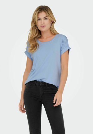 Donna con capelli lunghi e biondi che indossa una maglietta a maniche corte azzurro chiaro e jeans neri, in piedi davanti a uno sfondo bianco.