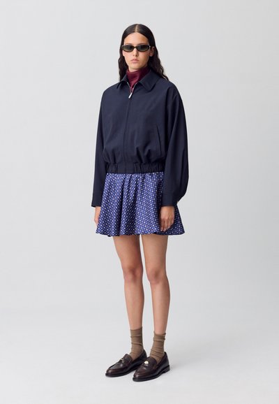 Chaqueta navy con cremallera, ajustada en la cintura, combinada con una falda acampanada que presenta un patrón de lunares azul y blanco; mocasines marrones y calcetines completan el look.