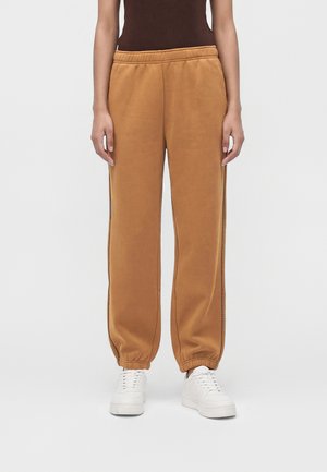 Persona que lleva pantalones jogger marrones con puños elásticos y zapatillas blancas, de pie frente a un fondo claro y liso.