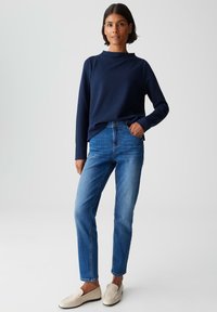 Femme debout portant un haut à manches longues bleu marine, un jean bleu et des mocassins blanc cassé, sur un fond clair uni.