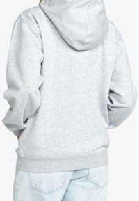 Personne portant un sweat à capuche gris clair et un jean en denim bleu clair, vue de dos avec les mains dans les poches.