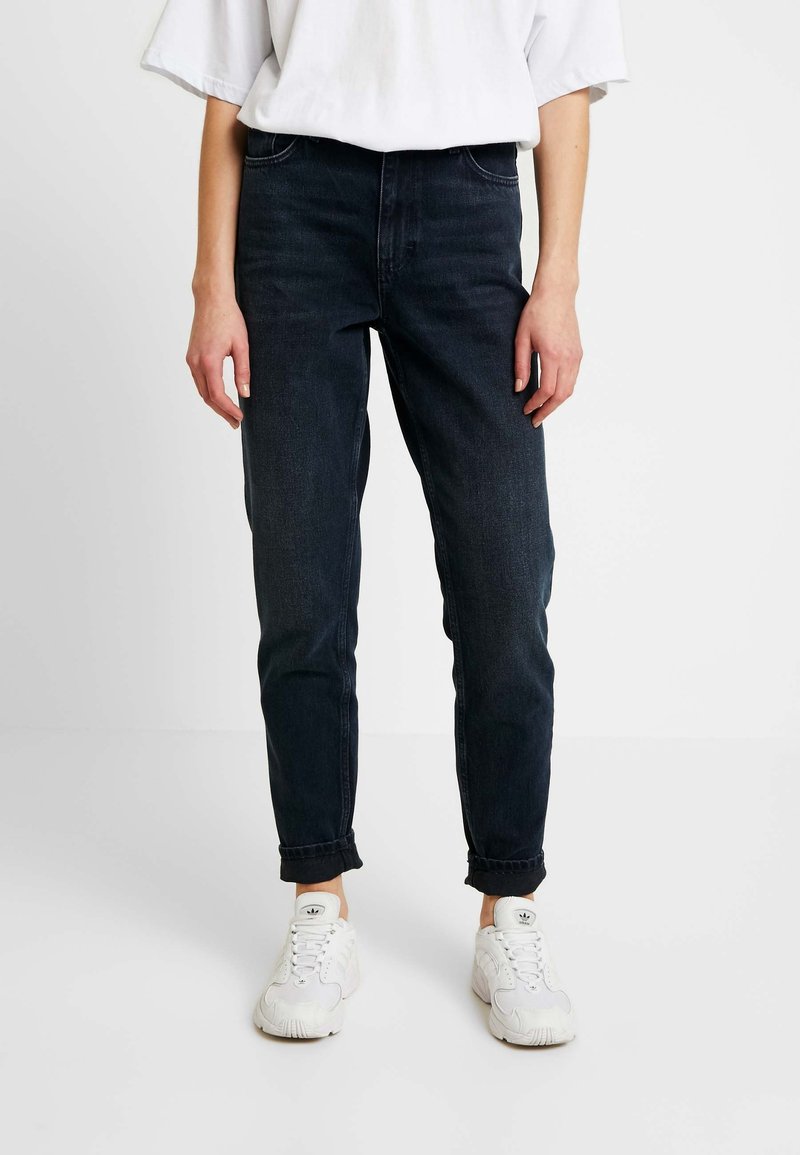 Jeans bleu foncé taille haute avec une jambe fuselée, dotés de cinq poches et de revers roulés, associés à des baskets blanches. Chemise en coton simple.