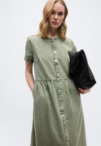 Γυναίκα με μαλλιά μέχρι τους ώμους που φοράει μια καλοκουμπωμένη ελιάς πράσινη κοντομάνικη φόρεμα, κρατώντας μια μεγάλη μαύρη υφή τσάντα φάκελο.