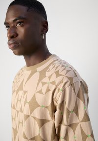 Beige langärmliges T-Shirt mit geometrischem Muster in hellbraunen und grünen Akzenten. Weiche Textur mit rundem Ausschnitt und lockerer Passform.