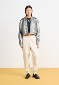 Srebrna skrajšana bomber jakna, črna majica s krožnim izrezom, kremna tapered hlače in črne loshle, stoječe na rumeni tla z enostavno ozadje.