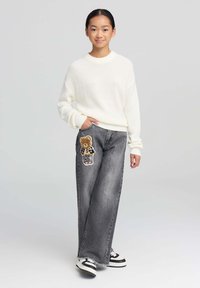 Przesadnie duży, kremowy sweter z dzianiny oraz luźne szare dżinsy z naszywką przedstawiającą niedźwiedzia na lewym udzie. Sneakery w czarno-białe paski.