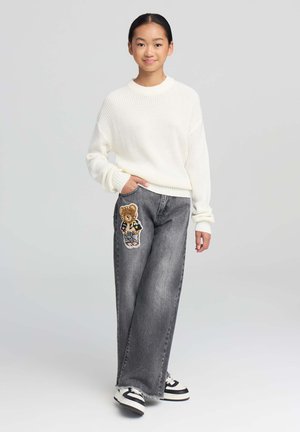 Maglione oversize beige a maglia e jeans in denim grigi larghi con una patch a forma di orso sulla coscia sinistra. Sneakers con strisce nere e bianche.