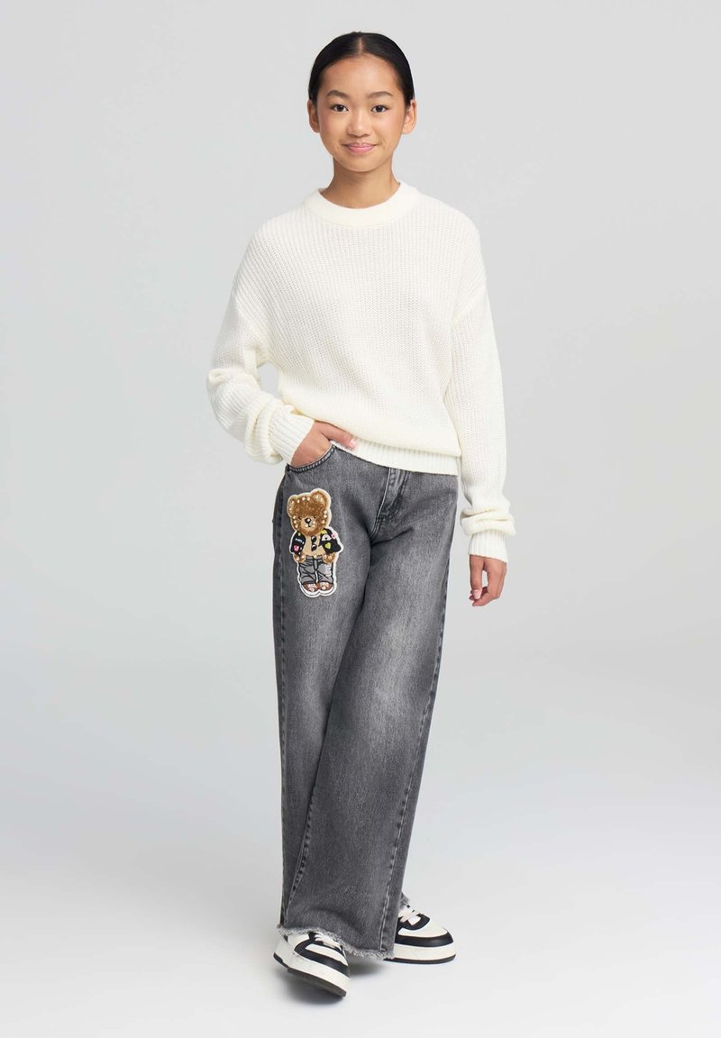 Przesadnie duży, kremowy sweter z dzianiny oraz luźne szare dżinsy z naszywką przedstawiającą niedźwiedzia na lewym udzie. Sneakery w czarno-białe paski.