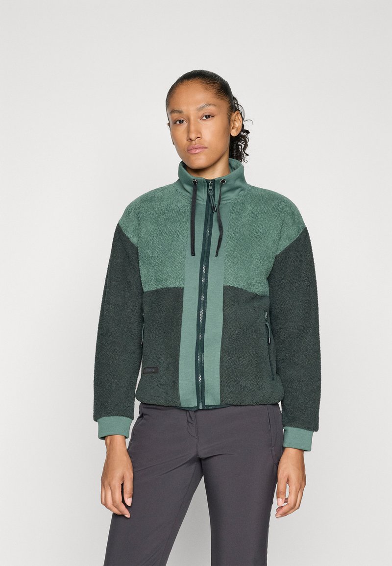 Woman wearing a green and dark gray zip-up fleece jacket with pockets and drawstrings, standing against a plain light gray background.
