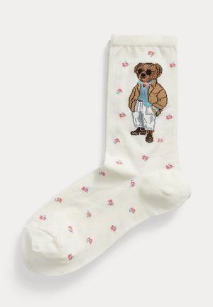 Calzino bianco con piccoli fiori rosa e blu e un orsetto vestito con occhiali da sole, giacca marrone, camicia blu e pantaloni bianchi.