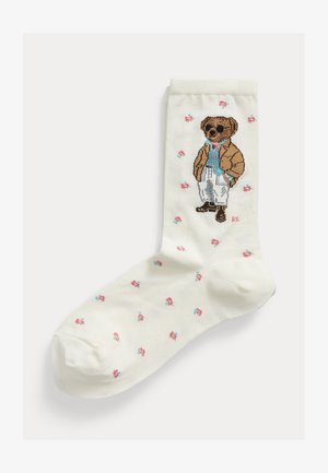 Calzino bianco con piccoli fiori rosa e blu e un orsetto vestito con occhiali da sole, giacca marrone, camicia blu e pantaloni bianchi.