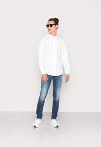 Witte lange mouwen overhemd met knopen, blauwe vervaagde jeans en witte sneakers. Accessoires zijn donkere zonnebril. Eenvoudig ontwerp en klassieke pasvorm.