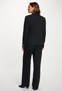 Mujer vestida con un traje negro de rayas diplomáticas con botones dorados, de pie en una pose de tres cuartos hacia atrás contra un fondo claro y sencillo.