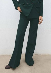 Tamnozelene krojene hlače s lepršavim nogavicama, izrađene od glatkog materijala, u kombinaciji s smeškim loafericama, prikazane na svijetloj pozadini.