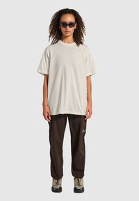 Lys beige oversized t-shirt, glat stof, rund halsudskæring. Kombineret med mørkebrune bukser med logo detalje. Modellen bærer sorte solbriller.