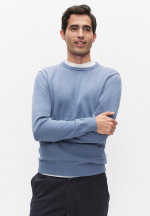 Jonge man met een lichtblauwe trui en donkere broek staat met gekruiste armen, kijkt iets naar de zijkant tegen een witte achtergrond.