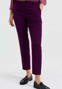 Sittande lila byxor med en slät textur, nedtill avsmalnande ben och sidofickor, kombinerat med svarta loafers för en välputsad look.