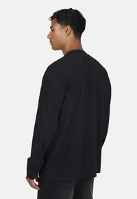 Man met kort, krullend haar, gekleed in een eenvoudige zwarte, lange mouwen shirt en donkere spijkerbroek, die naar links kijkt tegen een effen achtergrond.