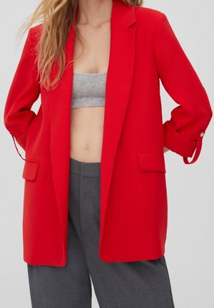 Woman wearing unbuttoned bright red blazer over gray crop top and dark gray high-waisted pants, hands behind back, light background.