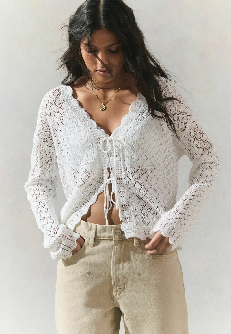 Cardigan en crochet blanc avec un col en V, fermeture nouée à l'avant, manches longues et bords festonnés. Associé à un pantalon taille haute beige clair.