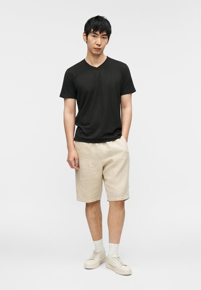 Μαύρο t-shirt με λαιμόκοψη σε σχήμα V, μπεζ λινά σορτς, λευκά αθλητικά παπούτσια και λευκές κάλτσες. Το μοντέλο στέκεται με τα χέρια στις τσέπες μπροστά σε απλό φόντο.